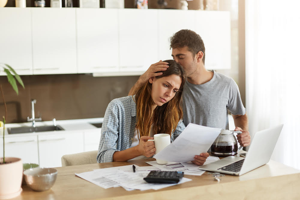 Frustrated woman studying sheet of paper in kitchen, drinking coffee and trying to find way to pay off all family debts; her husband kissing her on head